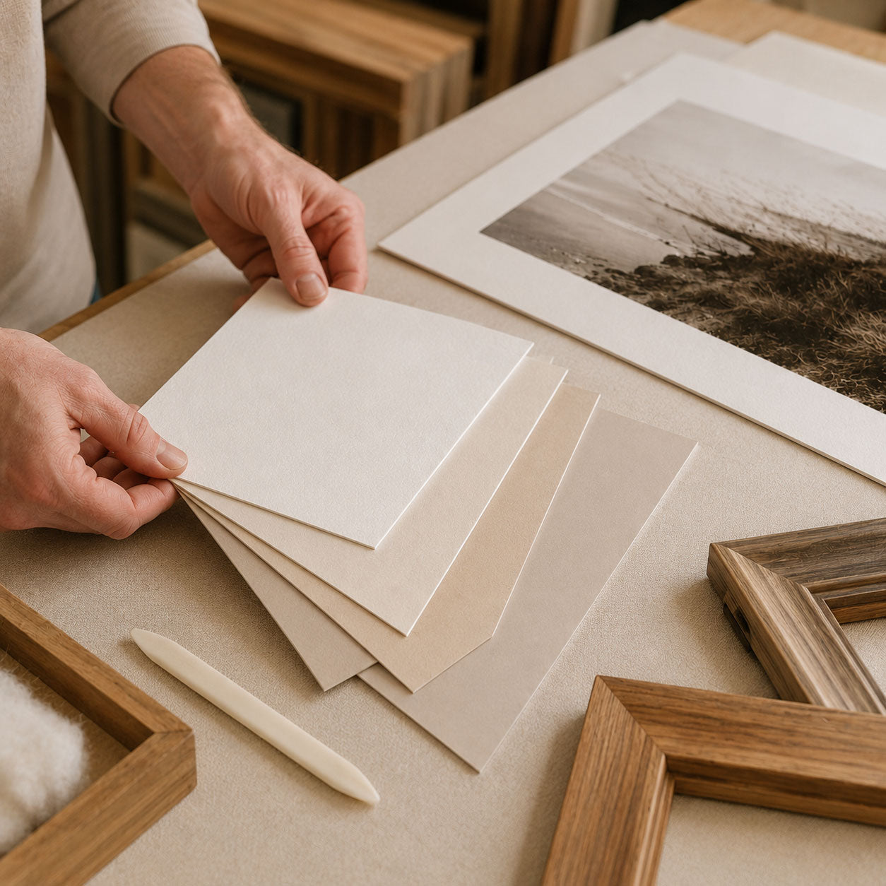 Hands selecting archival mat boards beside a framed print and wood moulding samples in a Germotte custom framing workspace.